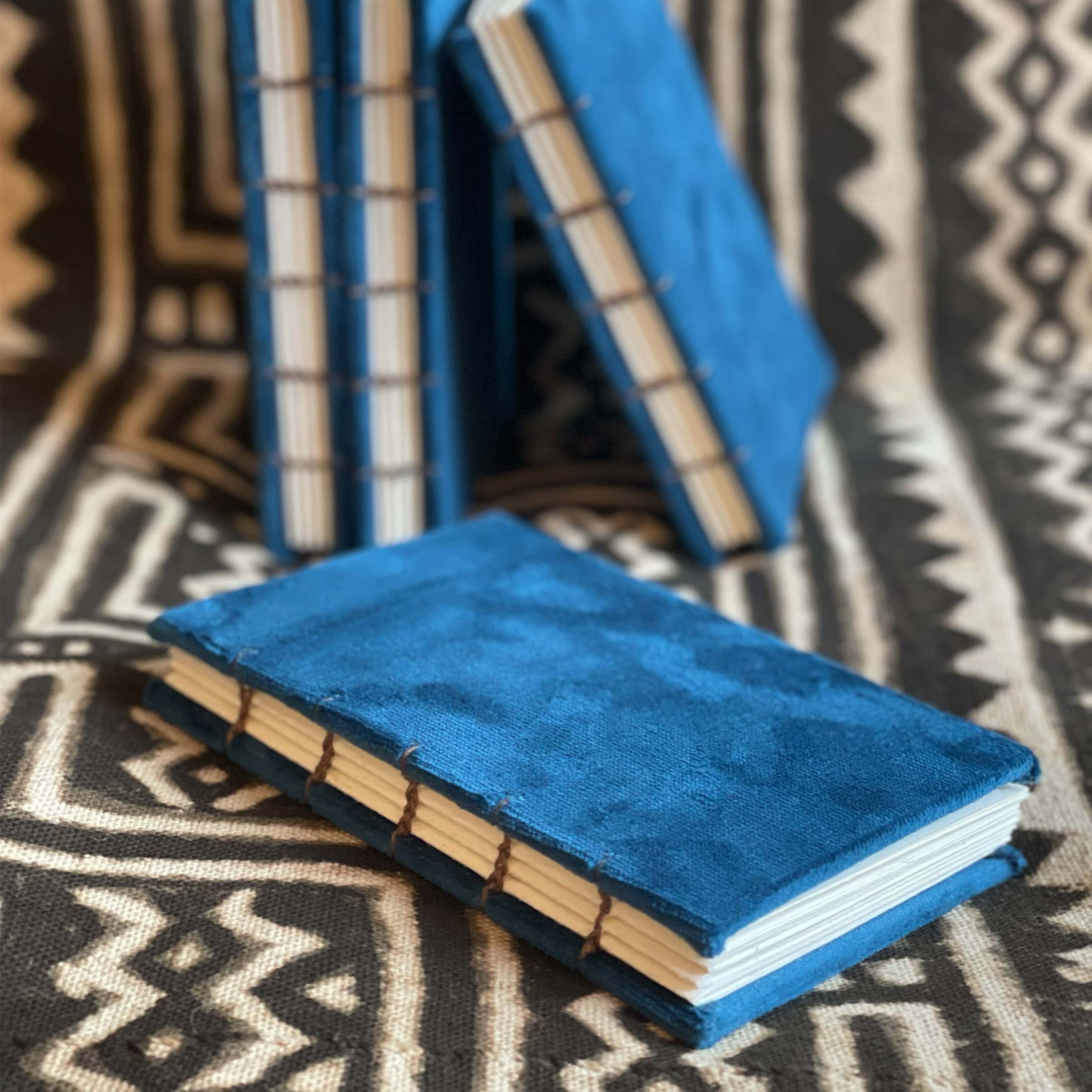 This is an image of handmade sketchbooks. This particular image features four of them standing upright against an African patterned blanket, with one laying down in front of the others. All four books feature a clear image of the binding